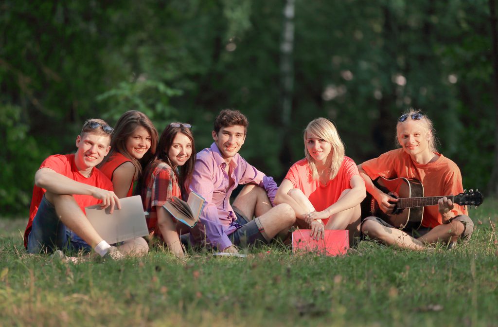 Grupa przyjaciół studentów z książakmi i gitarą siedzą na trawie w parku